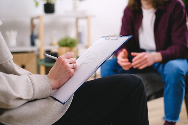 Person taking notes on a clipboard while talking with another seated individual in a casual office setting